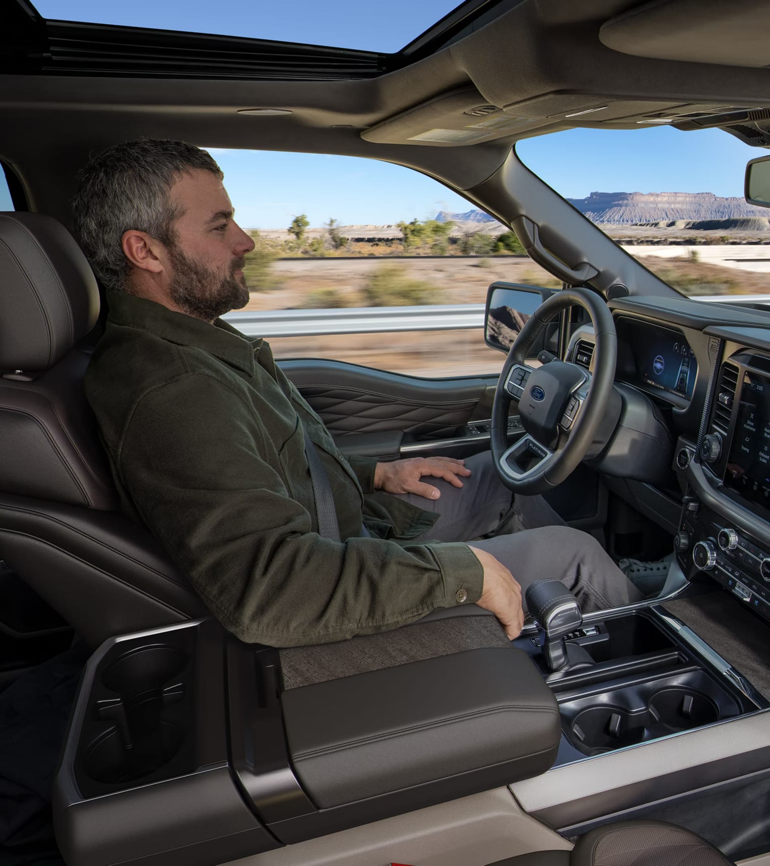 Person in a green jacket sitting in the driver's seat of a modern pickup truck, with a blurred desert landscape visible through the windows.