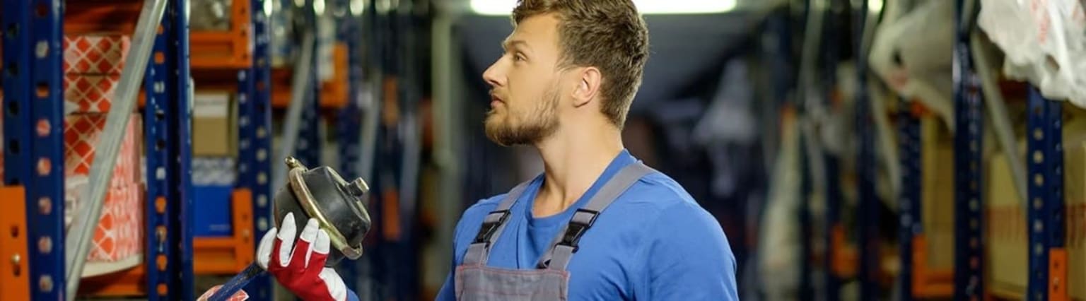 A man in blue shirt and overalls stands in a warehouse-like setting, surrounded by shelves and equipment.