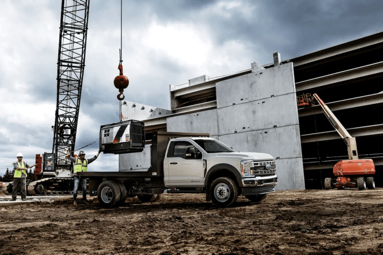 A white flatbed truck on a dirt construction site with a crane, workers in the foreground, a large concrete structure, and an orange boom lift in the background.