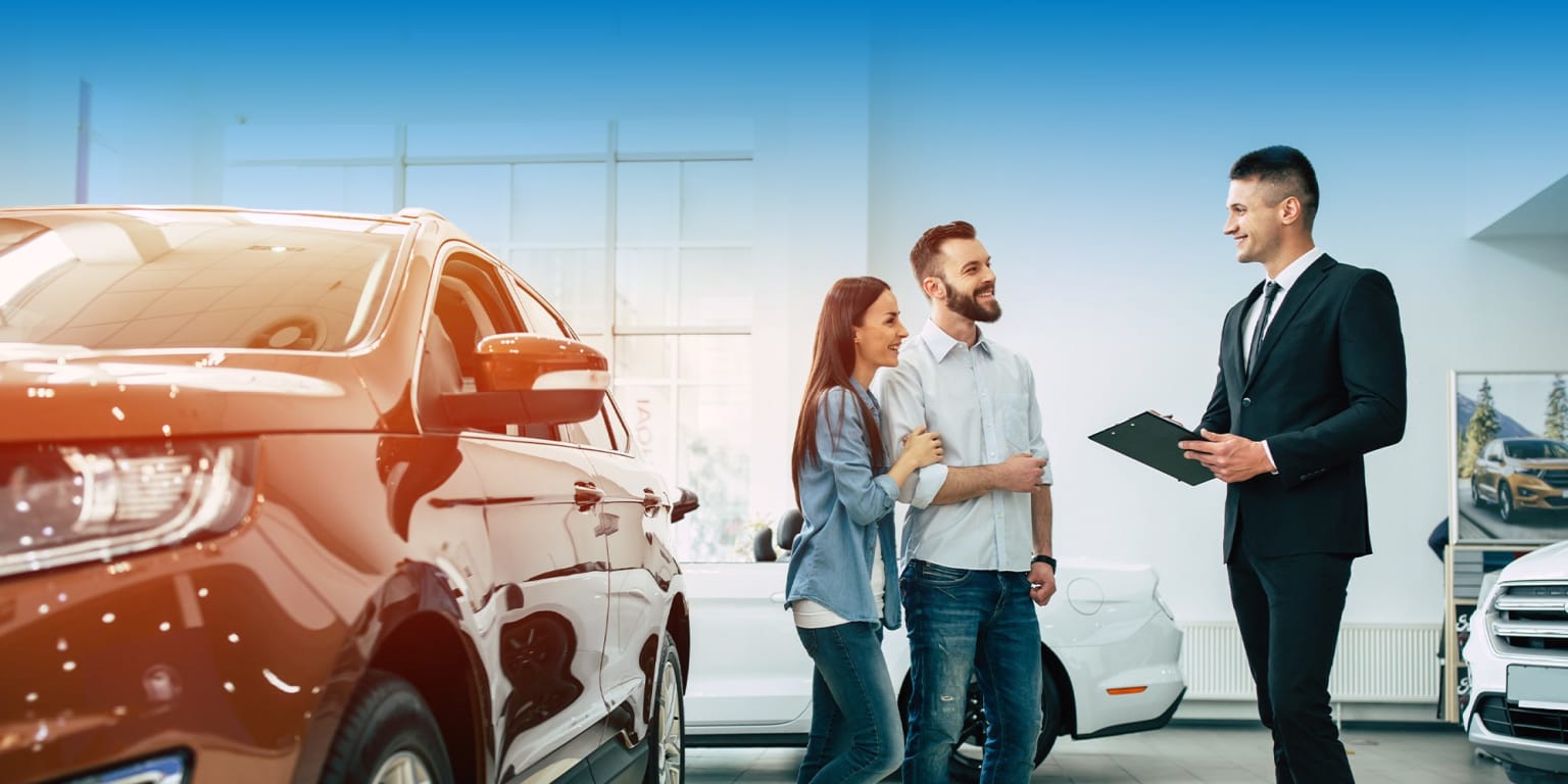 A couple with a car salesman in a brightly lit showroom, with a red car in the foreground and other vehicles and large windows in the background.