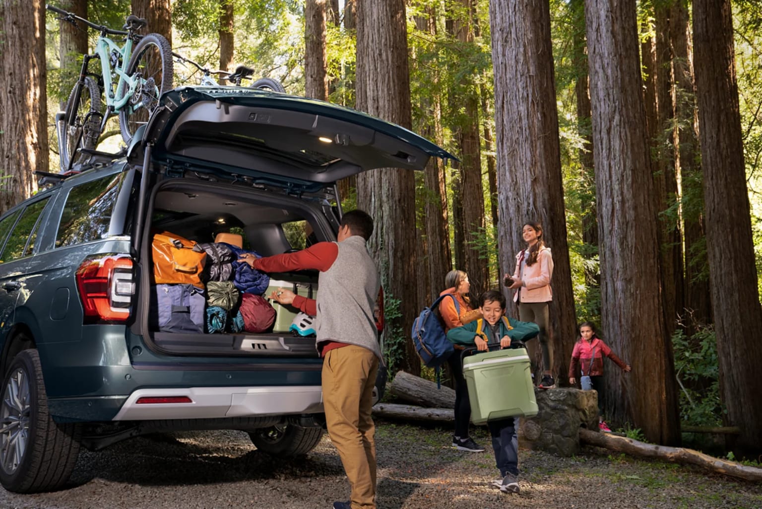 A group of people, including children, are gathered around an open vehicle in a lush, forested environment, suggesting an outdoor adventure or camping trip.