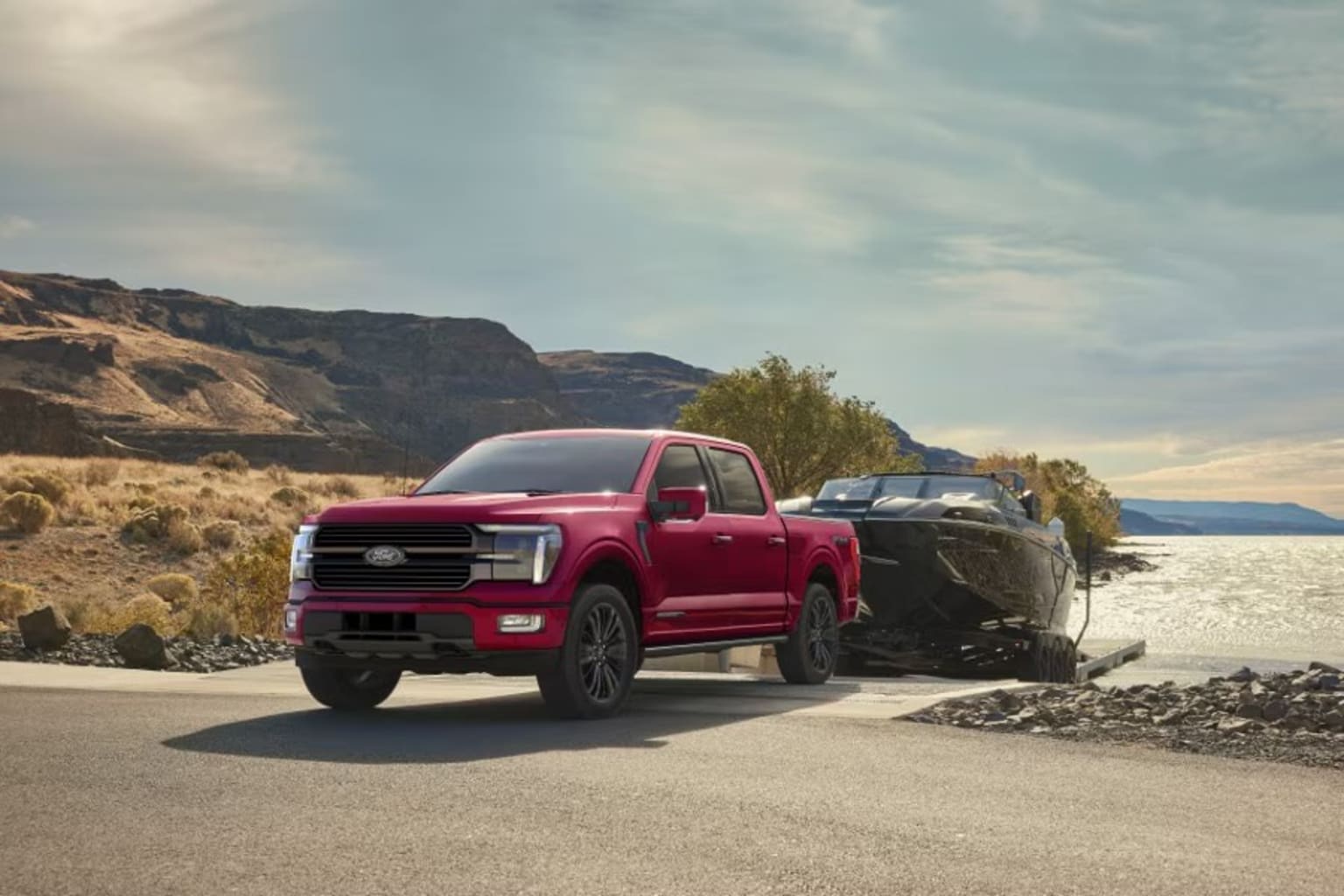 A red pickup truck is parked on a dirt road, with rugged mountains and a cloudy sky in the background.