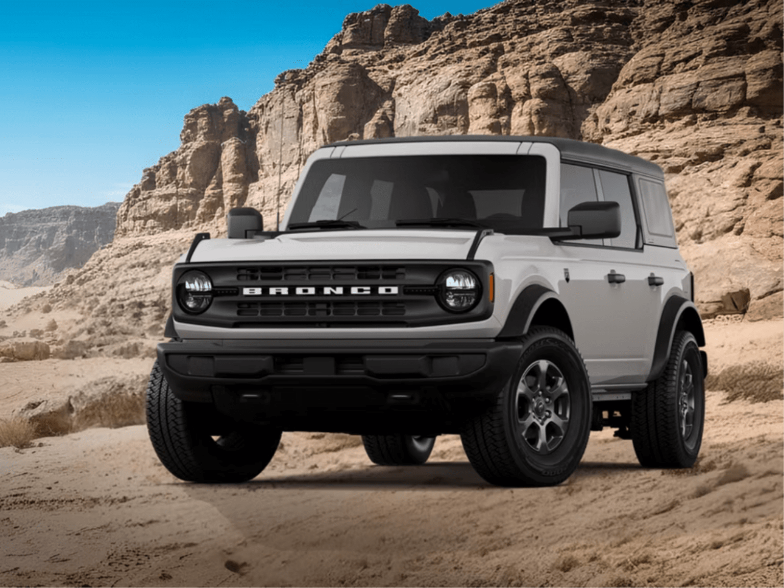 A white SUV sits on a sandy desert floor against a backdrop of towering red rock cliffs and a clear blue sky.