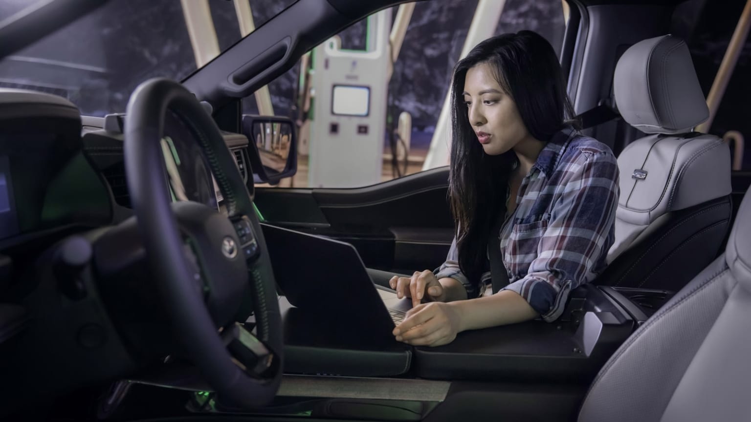 a woman sitting in the driver's seat of a car using a laptop computer while holding a cell phone