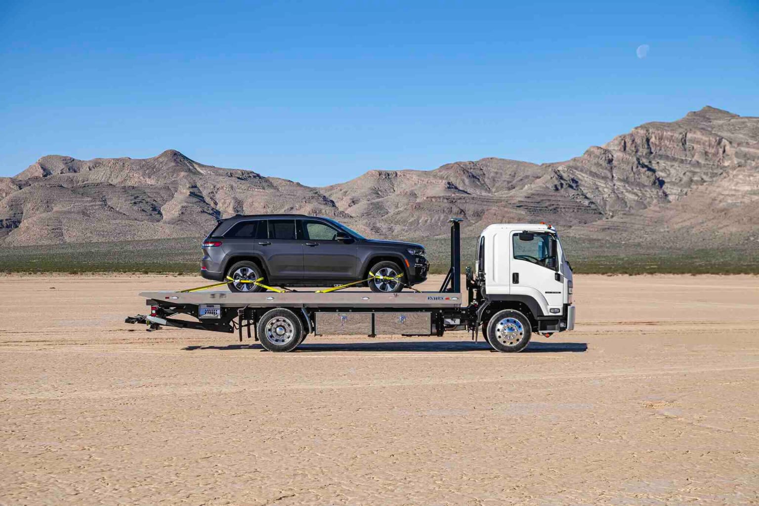 A gray SUV on a flatbed tow truck in a desert landscape with mountains in the background.