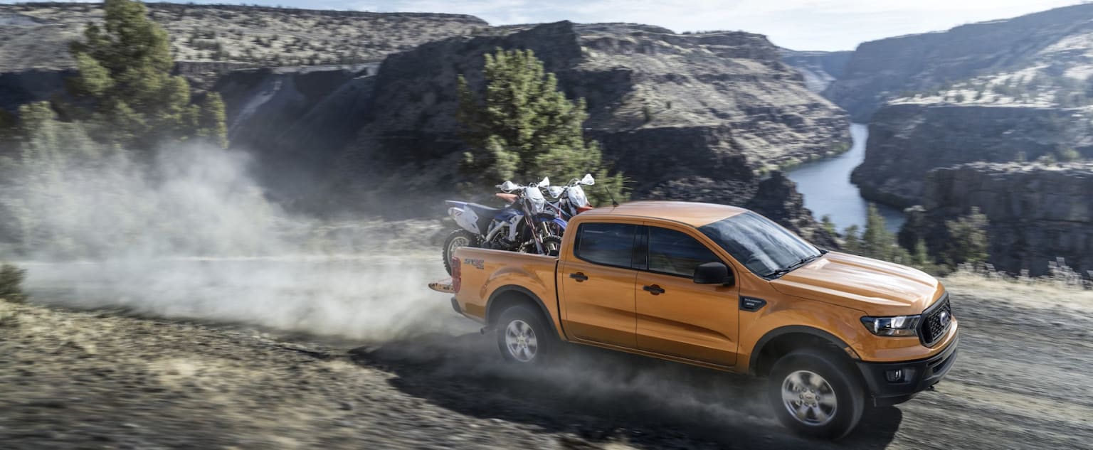 an orange pick up truck driving down a dirt road near a lake and a mountain range with a motorcycle on the back of the truck