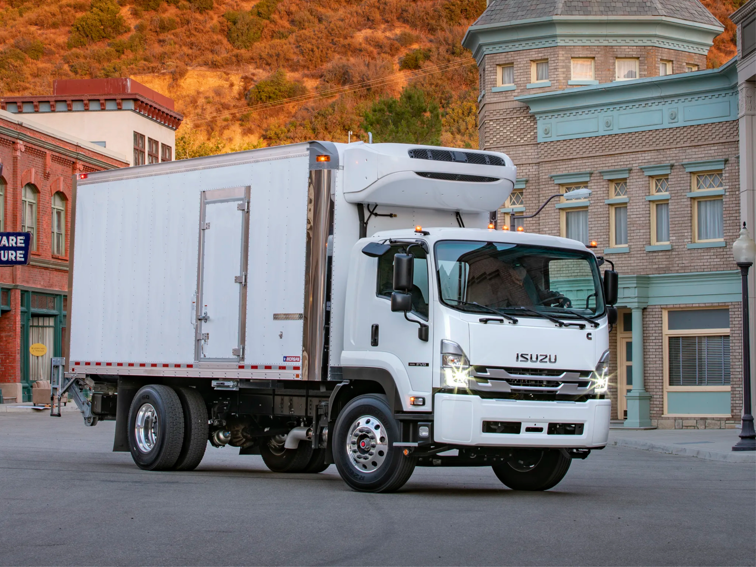 A white box truck is parked on a city street in front of historic buildings and a sunlit hillside.