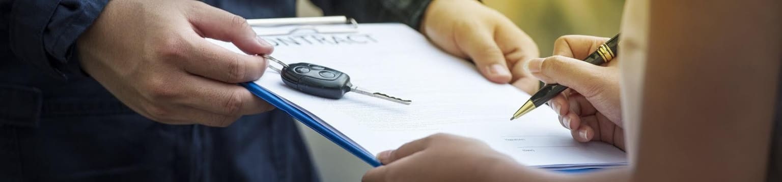 An image of a person's hands holding a car key and a pen, signing a document on a desk.
