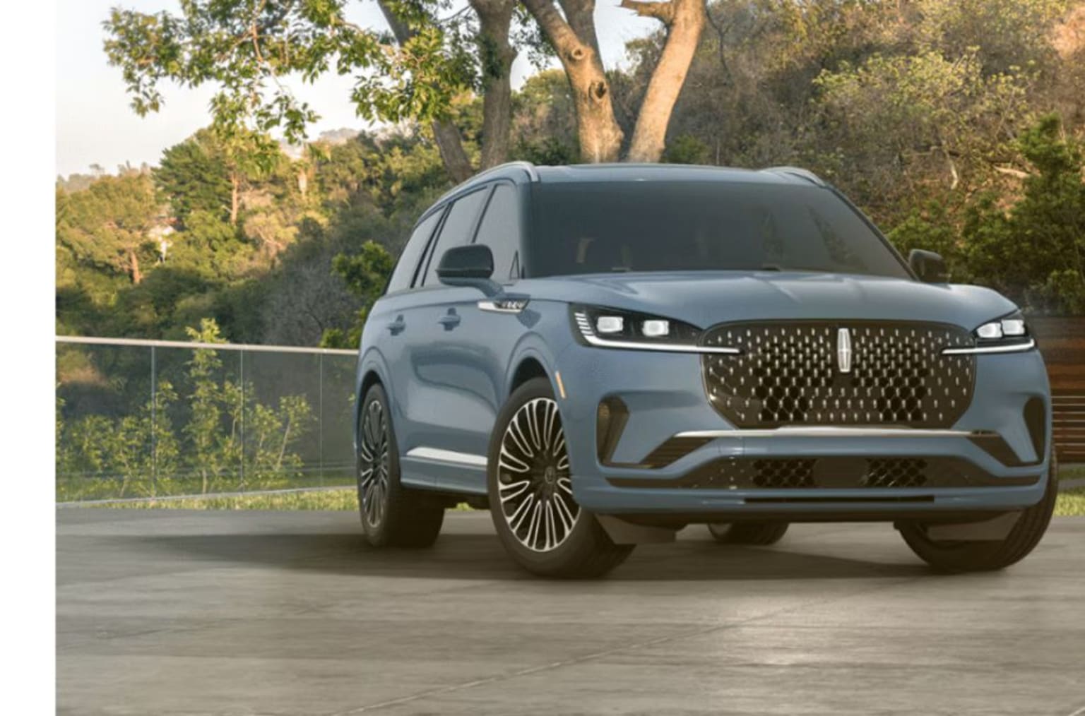 A large, sleek gray SUV with a distinctive grille design parked on a paved surface, surrounded by lush greenery and trees in the background.