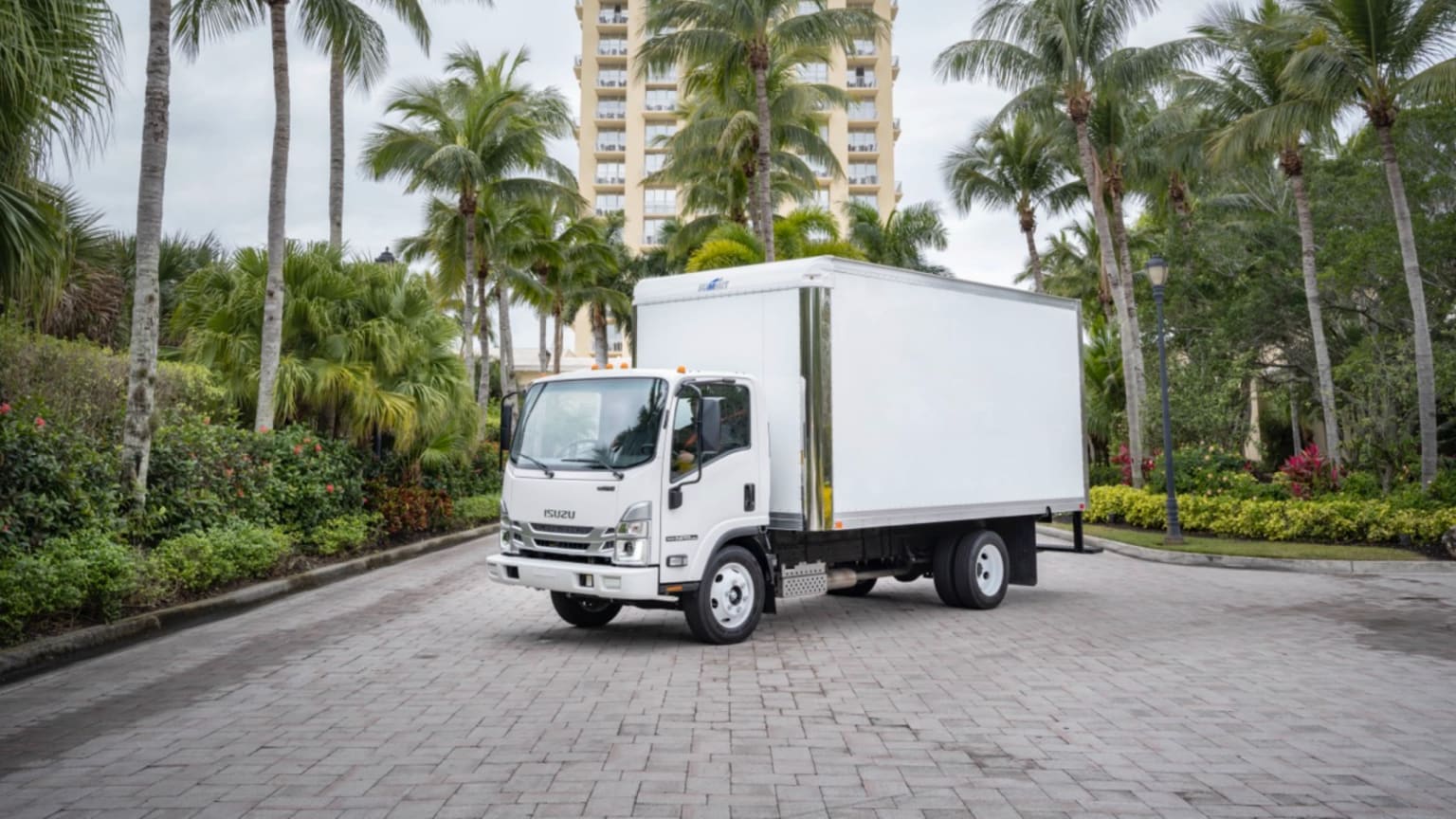 A delivery truck is parked on a brick-paved road, surrounded by lush palm trees and tall buildings in a tropical setting.
