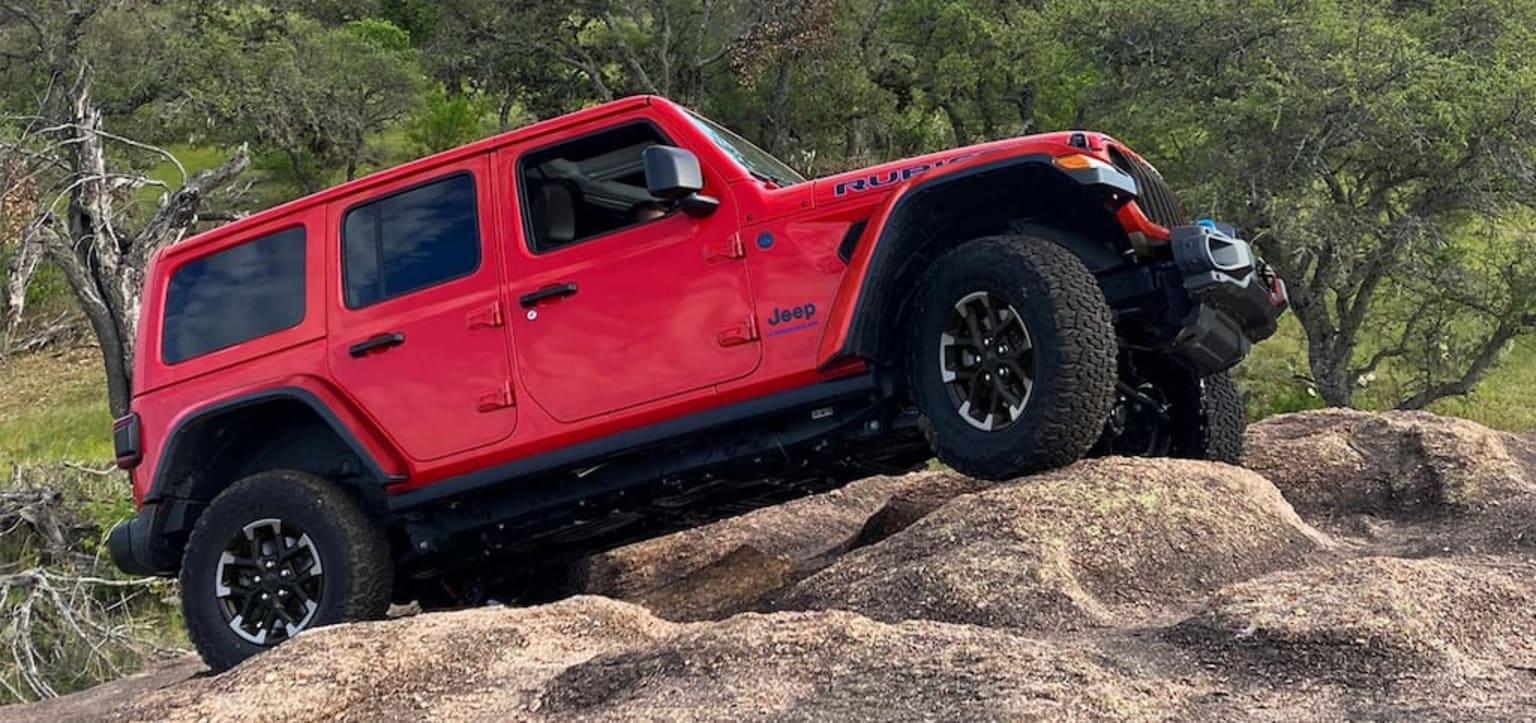 The image shows a red off-road vehicle navigating over rocky terrain in a forested area with lush greenery in the background.
