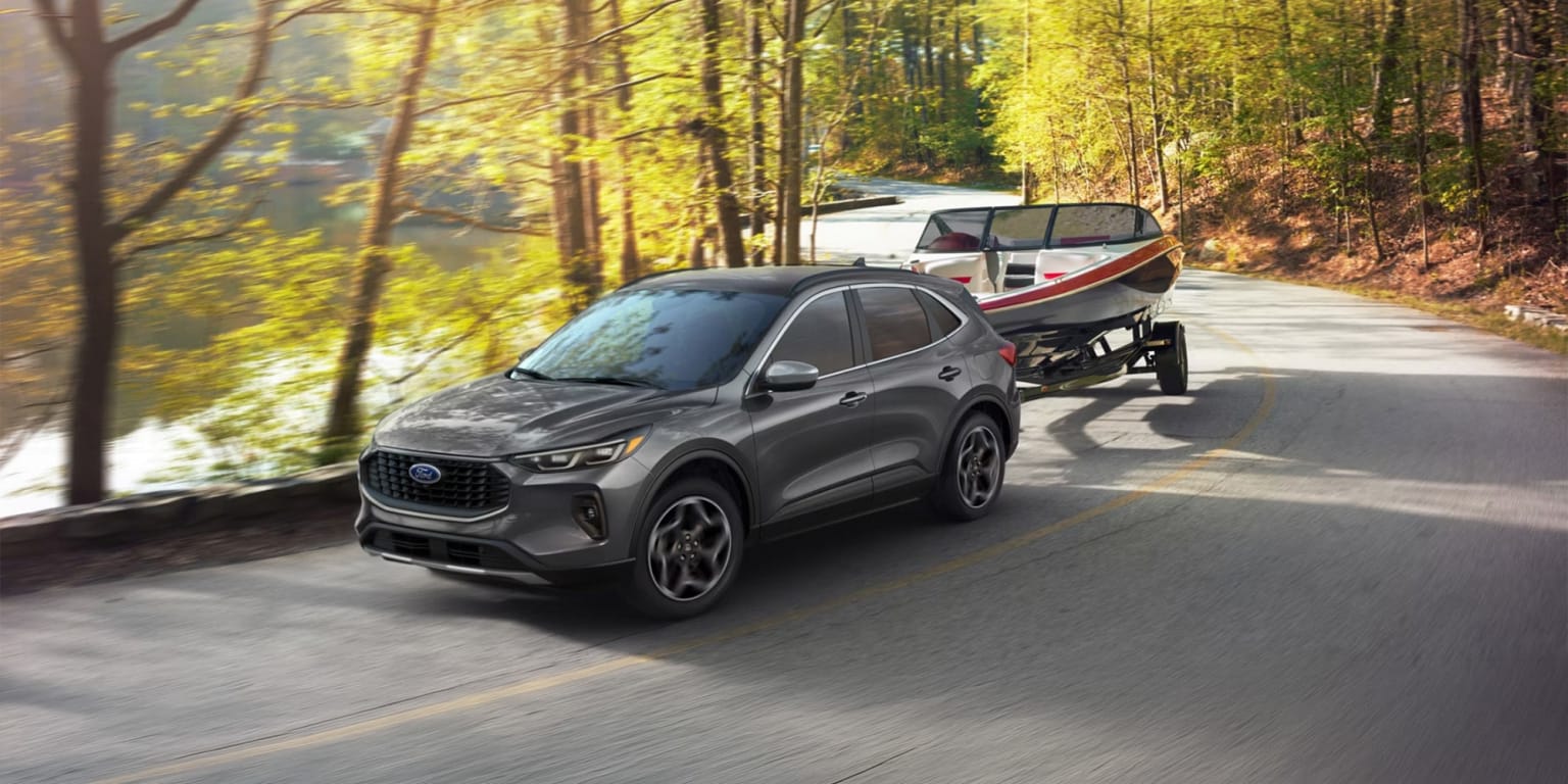 A gray SUV driving down a winding road surrounded by a lush, autumnal forest with sunlight filtering through the trees.