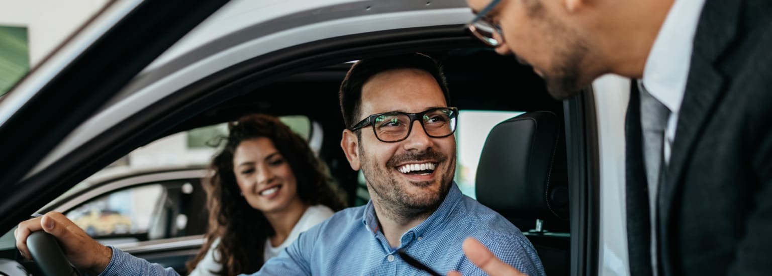 A man in glasses sits in the driver's seat of a car with a woman behind him, as a salesman in a suit talks to them in a dealership.
