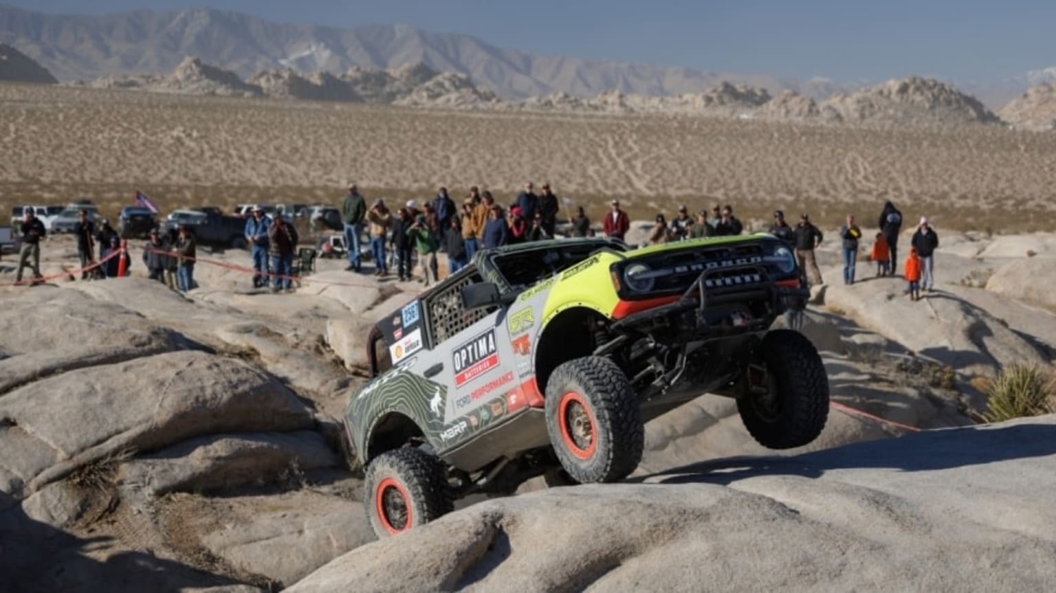 a monster truck is jumping over rocks in the desert with a crowd of people watching from the side of the road