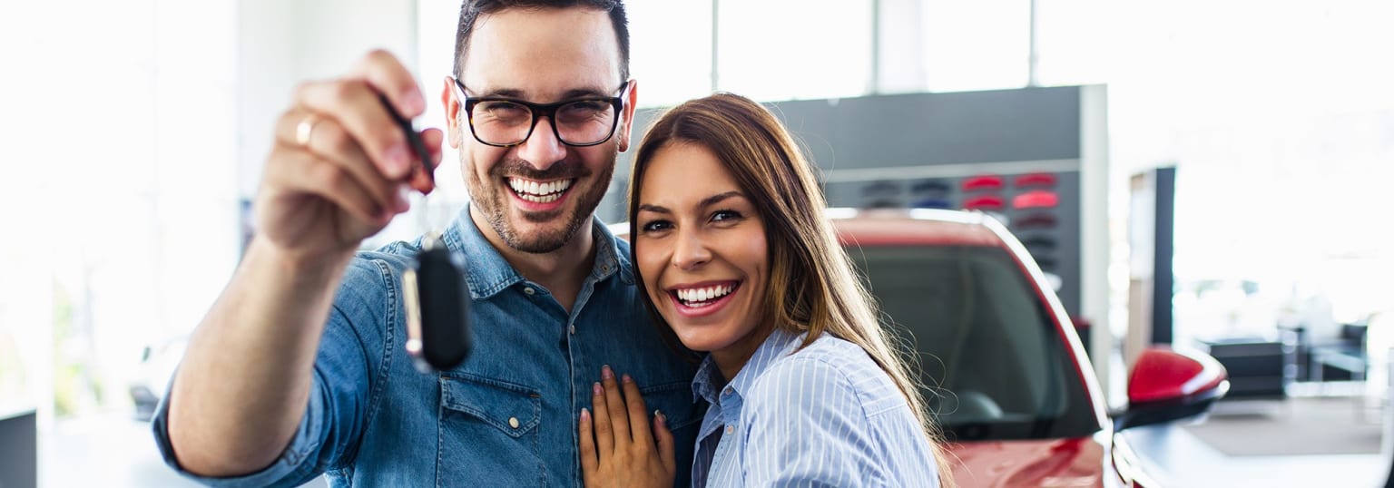 A smiling couple, a man wearing glasses and a denim jacket and a woman with long hair, posing together in a car showroom with vehicles visible in the background.