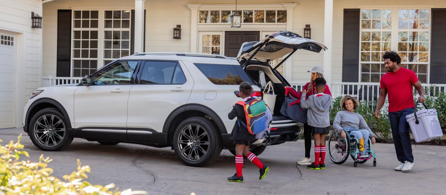 A family is loading their vehicle with luggage and sports equipment, with a large white SUV parked in the driveway of a residential home in the background.
