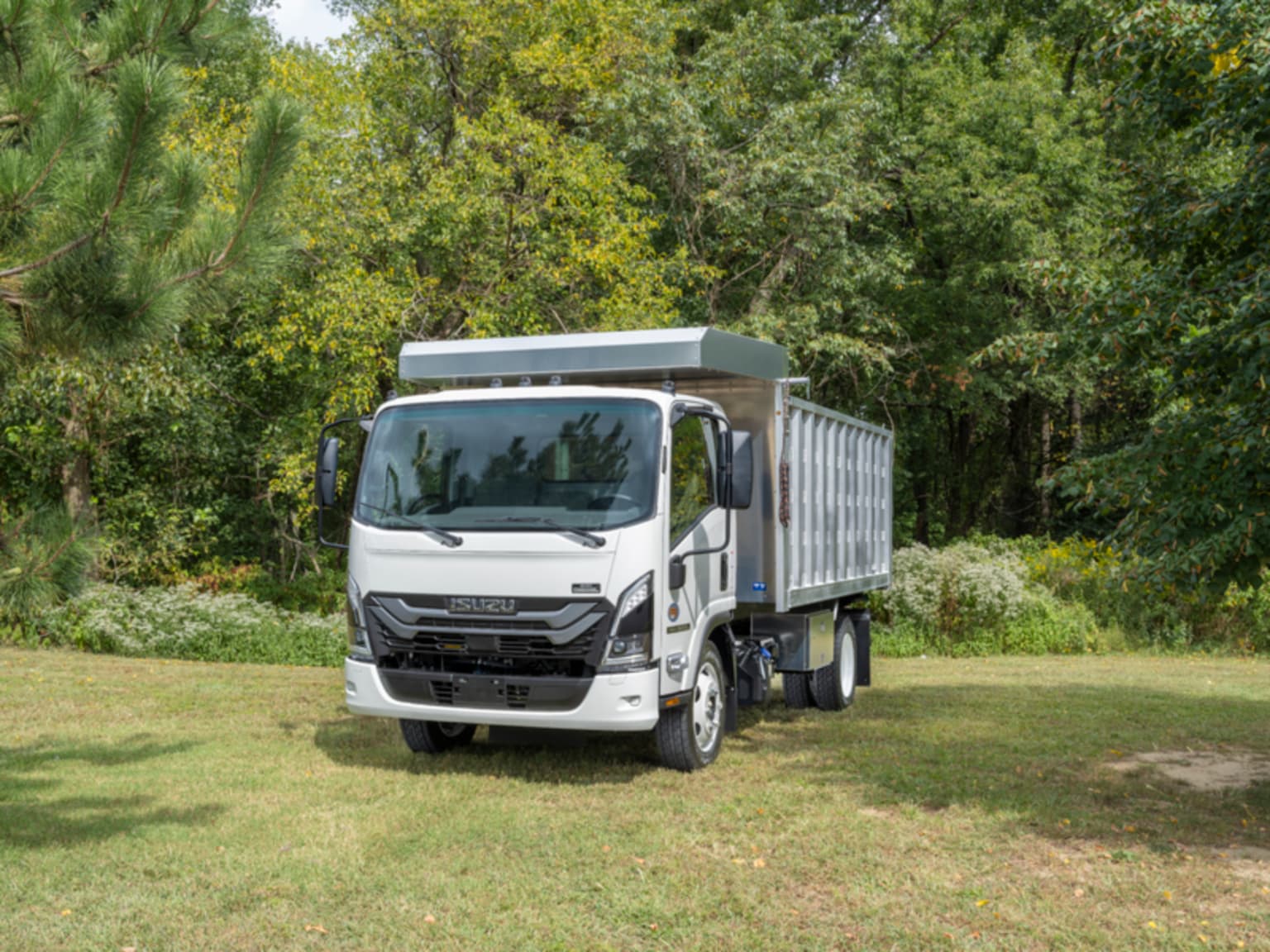 A white dump truck sits on a grassy field against a backdrop of lush green trees.