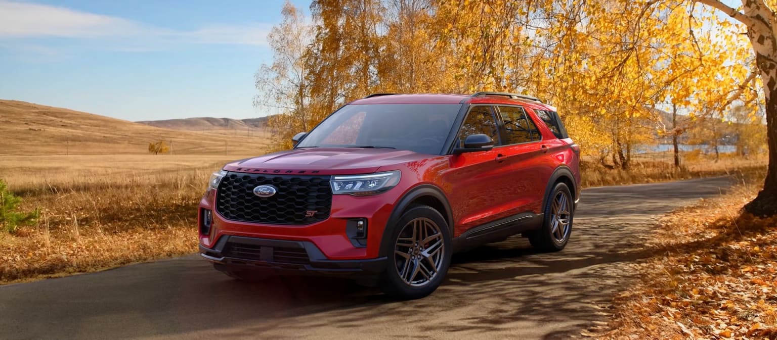 A bright red SUV with a bold grille design is parked on a dirt road, surrounded by a golden autumn landscape with a clear blue sky.