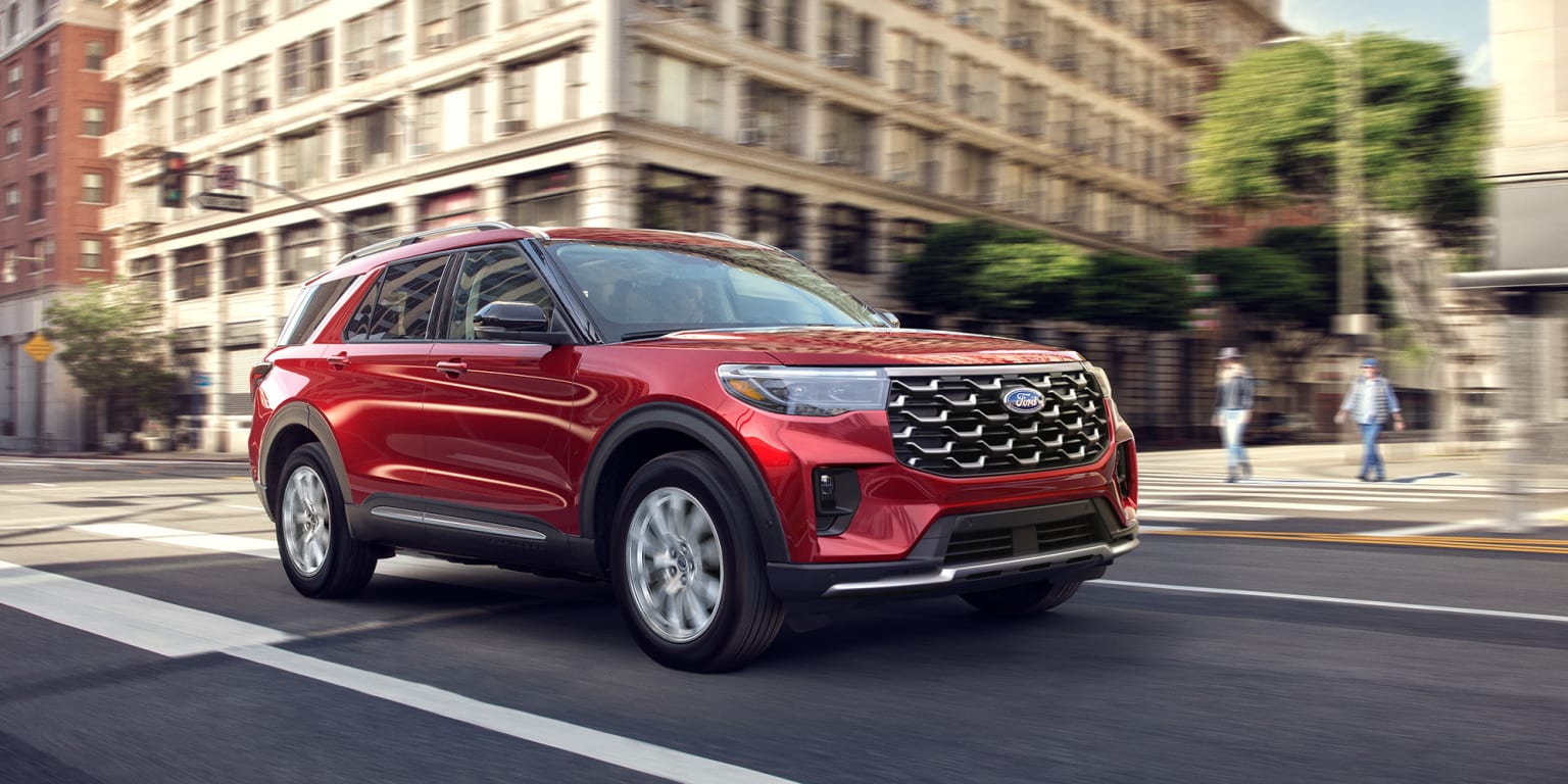 A red SUV is driving down a city street, with tall buildings and pedestrians visible in the background.