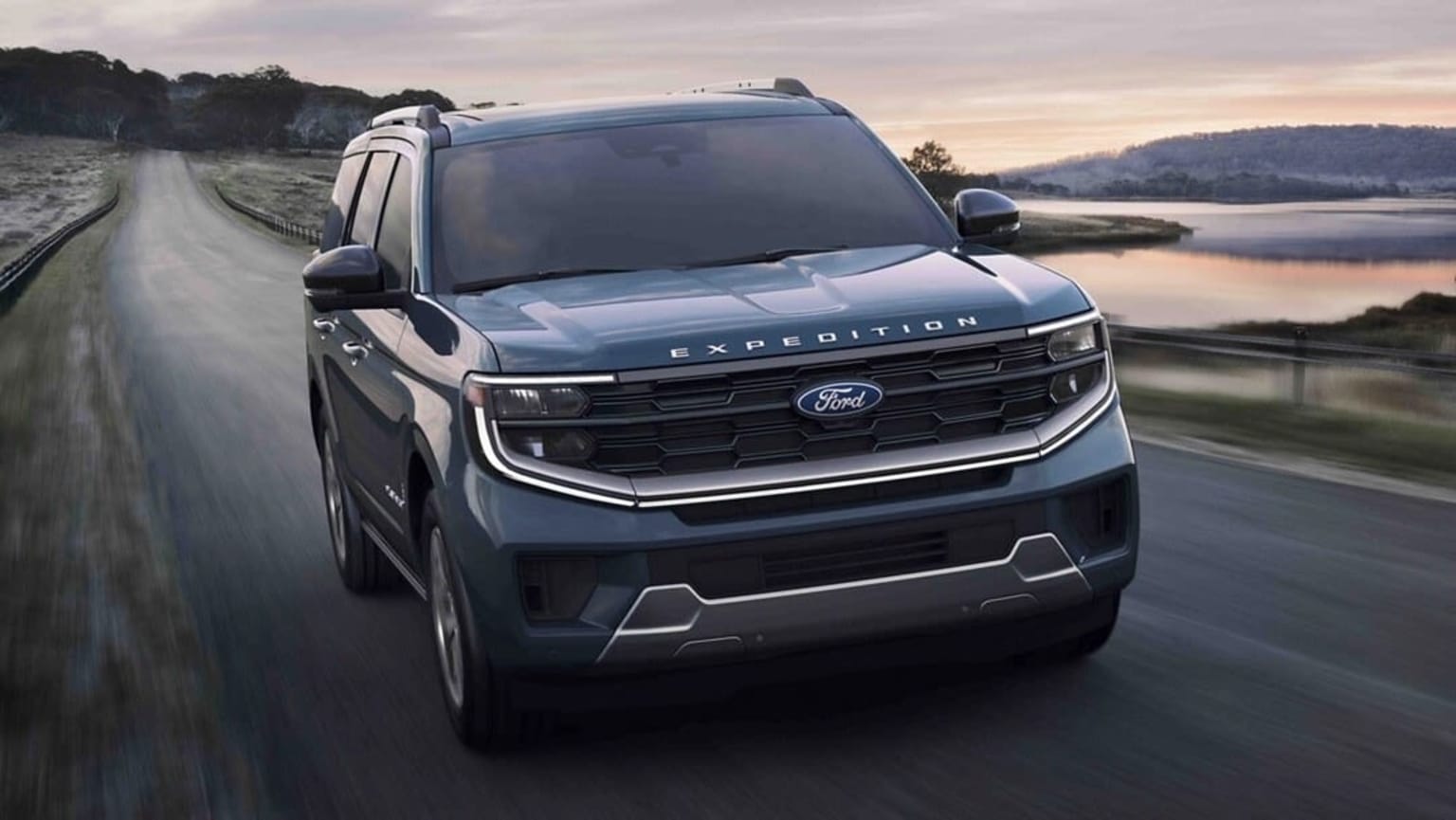 A dark blue SUV drives down a winding road with a calm body of water and hills in the background.