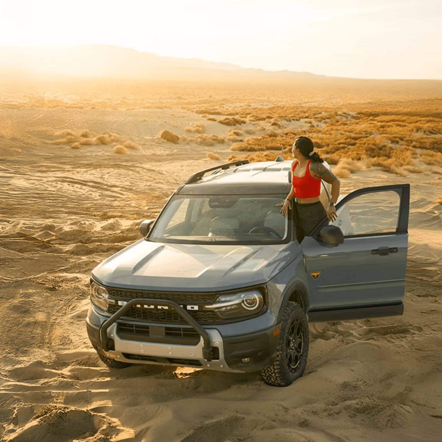 A woman in a red top leans against a gray SUV parked on sandy dunes, with a hazy, sunlit desert landscape stretching into the background.