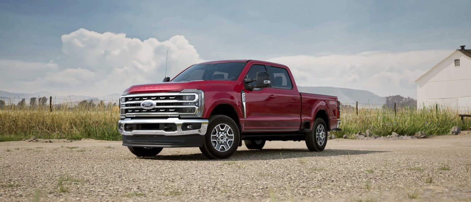A red pickup truck in a grassy field with mountains and cloudy skies in the background.