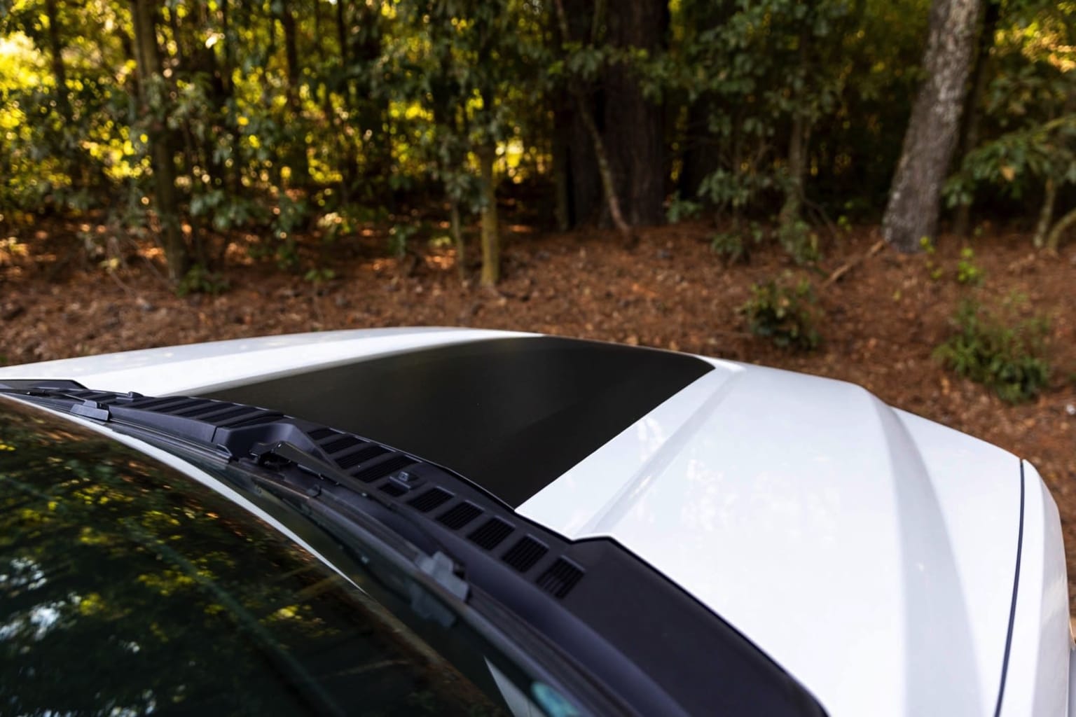 A white car with a sleek, angular design is parked in a lush, forested area, with sunlight filtering through the trees and casting a warm, golden glow on the scene.