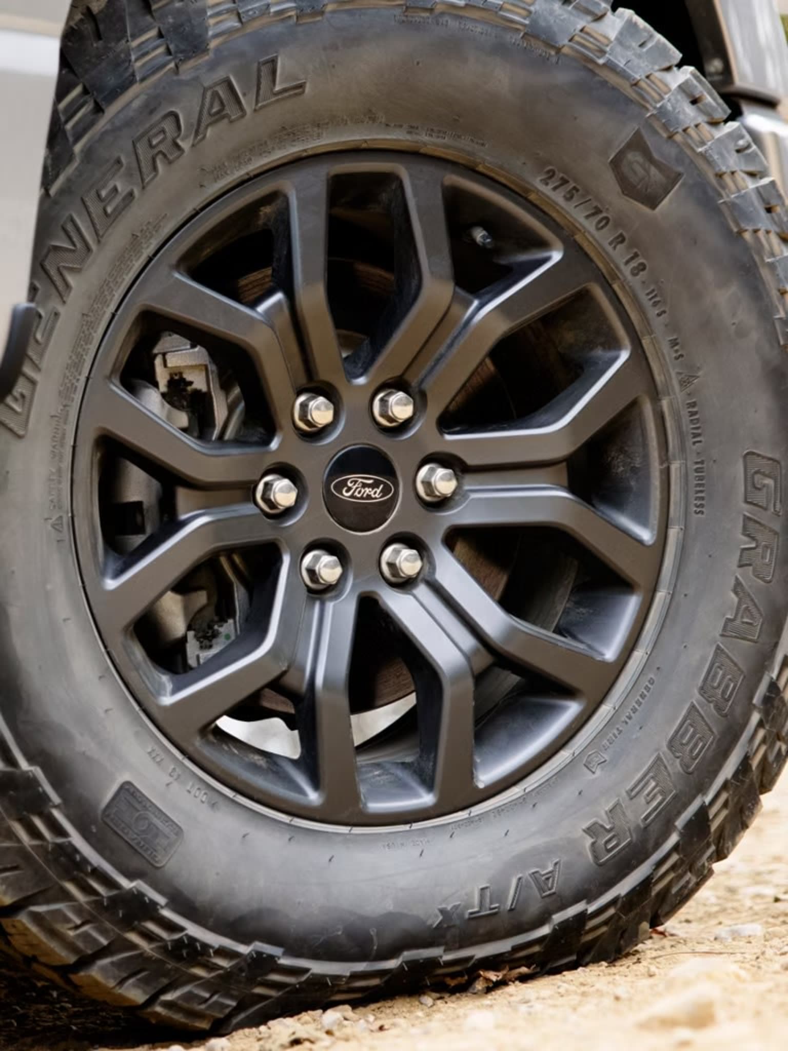 Close-up of a dark gray wheel with a General Grabber A/TX tire, with a blurred background of a light-colored vehicle and dirt.