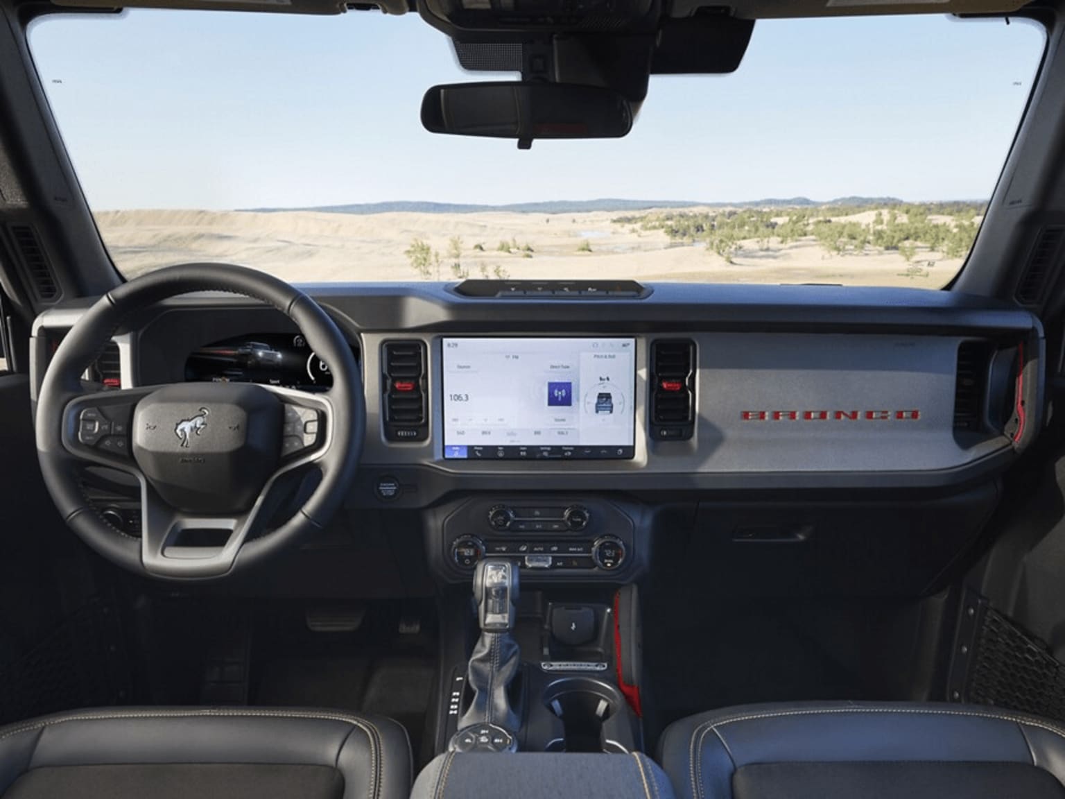 Interior of a vehicle with a steering wheel, dashboard, and a large touchscreen display in the center. Desert landscape visible through windshield.