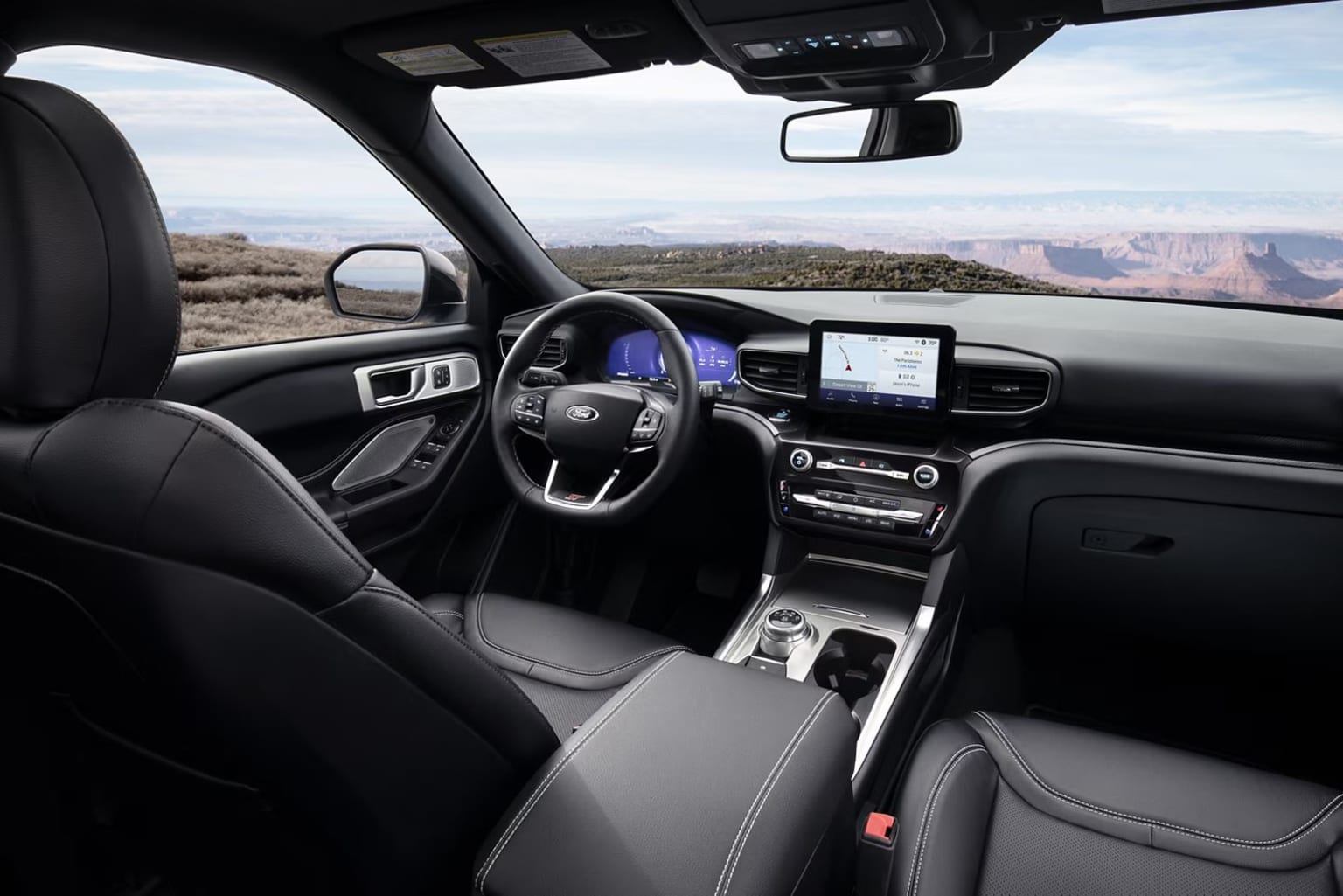 Interior of a modern vehicle with a large touchscreen display, steering wheel, and dashboard controls, overlooking a scenic desert landscape in the background.