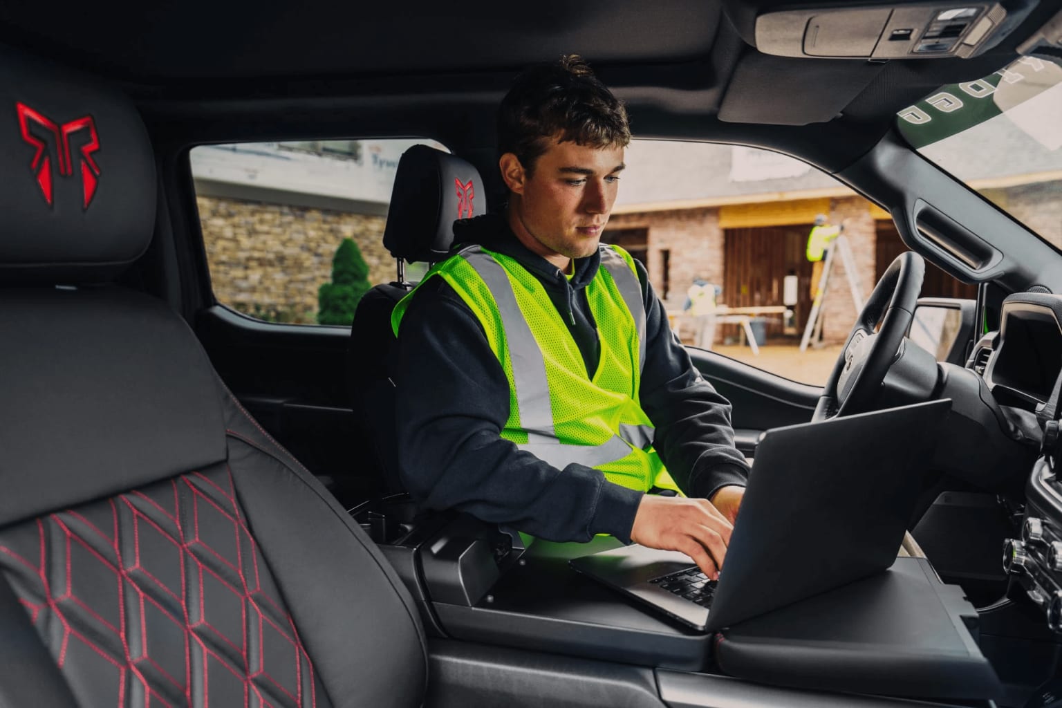 A person wearing a high-visibility vest is sitting in the driver's seat of a vehicle, using a laptop computer.