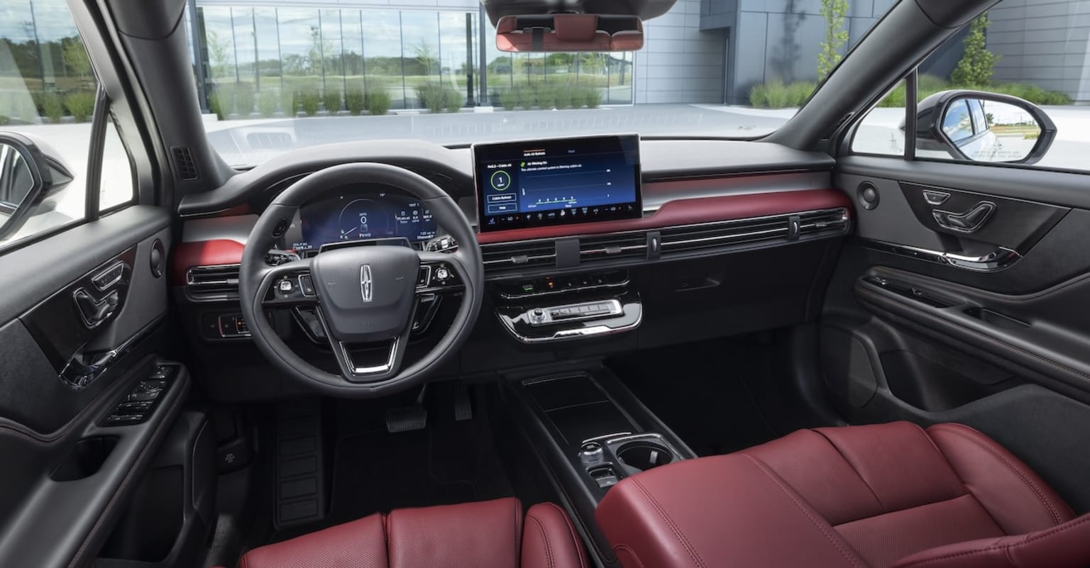 Interior of a modern vehicle with a sleek dashboard, large touchscreen display, leather-wrapped steering wheel, and red upholstery, against a backdrop of glass windows and a cityscape outside.