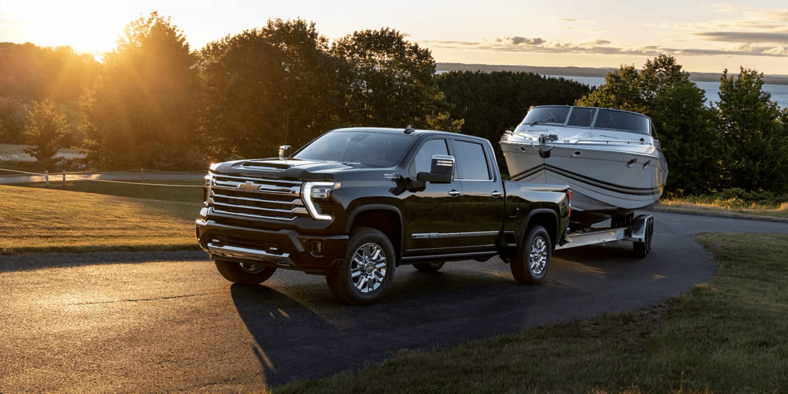 A pickup truck towing a boat trailer parked on a dirt road surrounded by green trees and a scenic landscape with the sunset in the background.