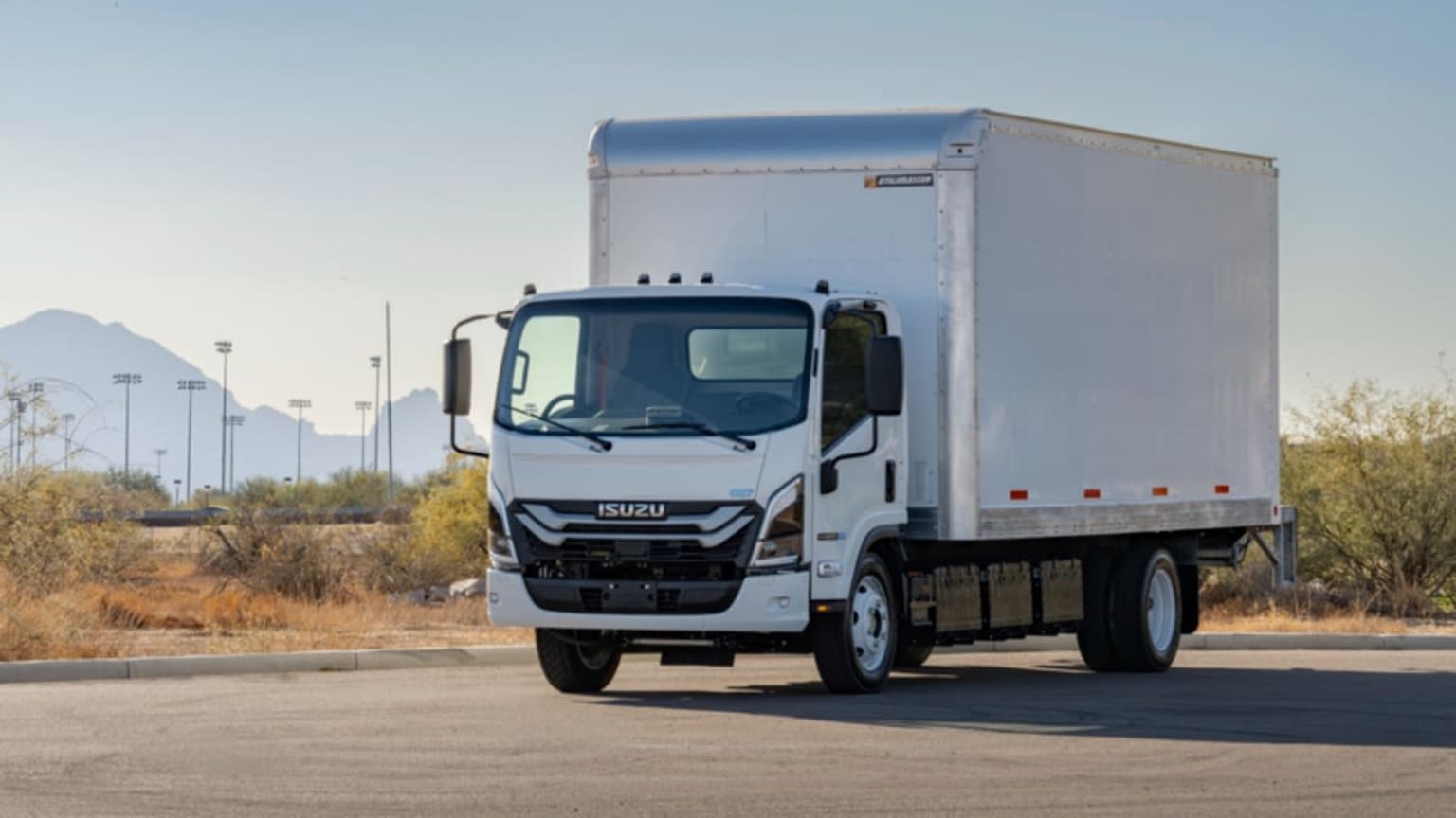 A white box truck is parked on a paved lot with dry brush and distant mountains under a clear sky.