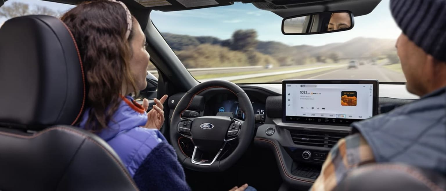 Interior view of a car with a driver looking through the windshield at the road and landscape. The person in the driver's seat is interacting with a touchscreen display on the center console.