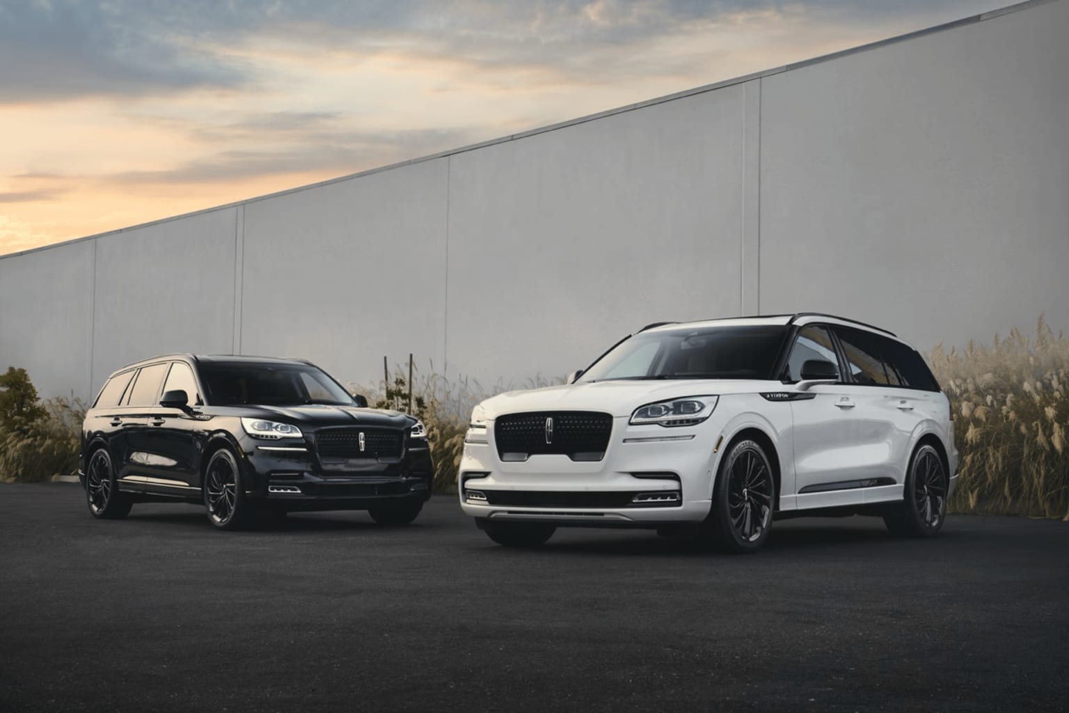 Two luxury SUVs, one black and one white, parked in front of a gray concrete wall against a cloudy sky at sunset.