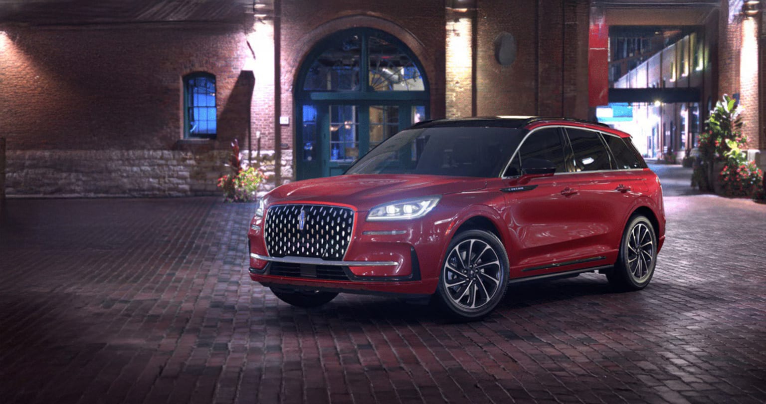 A large, red luxury SUV is parked on a cobblestone street, with a dimly lit building and storefront visible in the background.