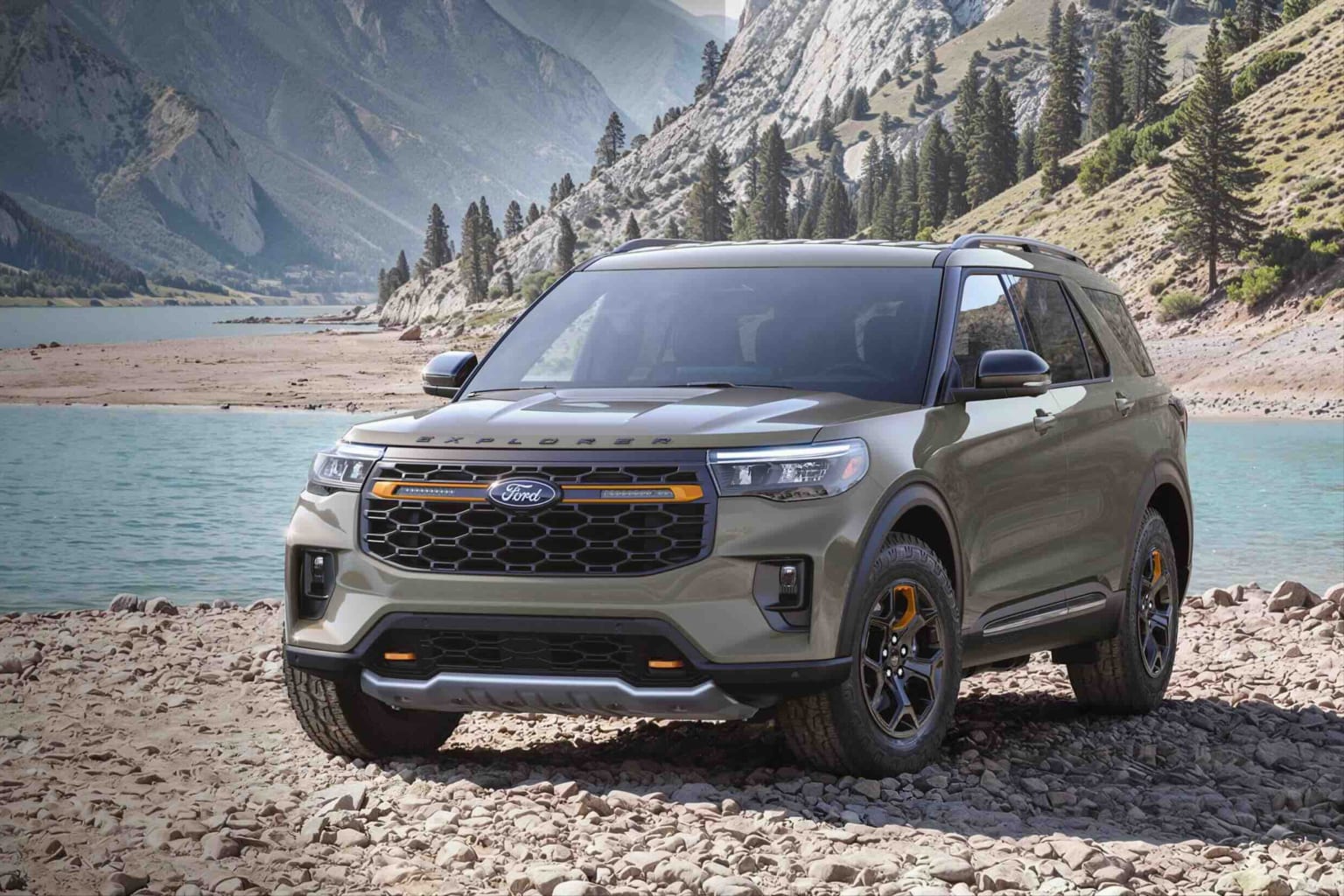 An olive-green SUV parked on a rocky shore with a calm lake and mountainous landscape in the background.