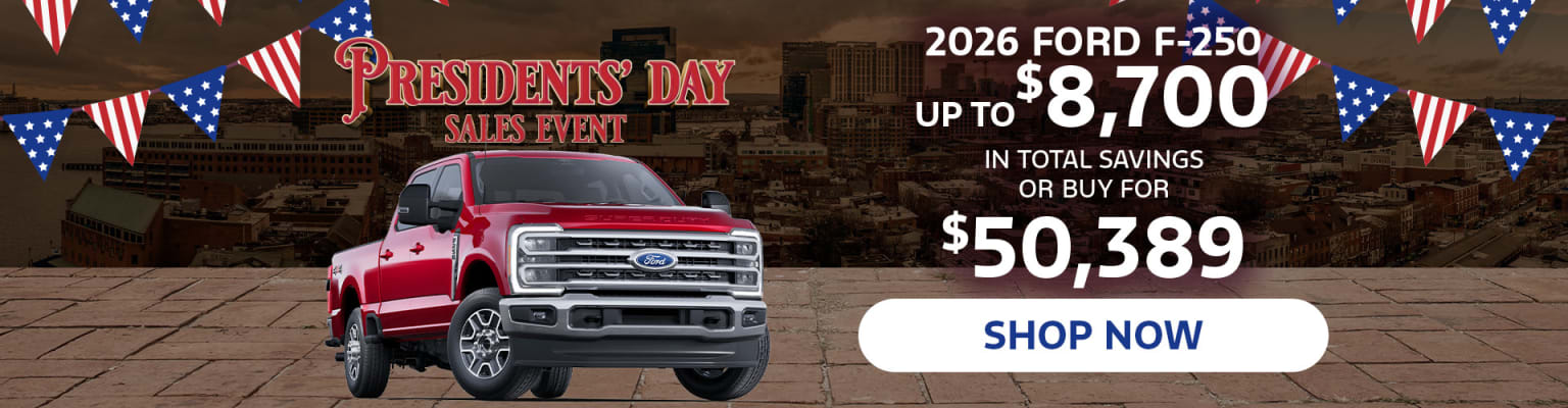 A large red pickup truck displayed in the foreground, with a Presidents' Day sales event advertisement in the background showcasing discounts for the 2026 model year.