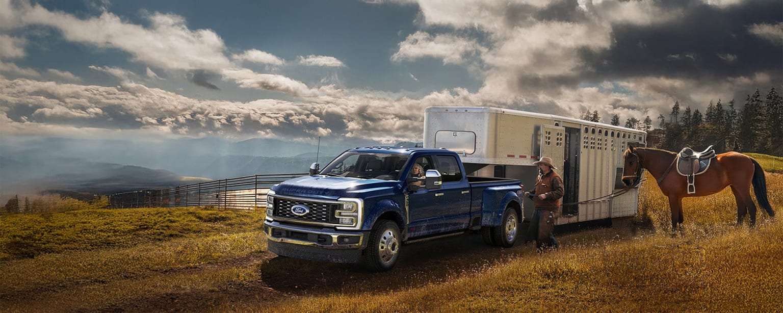 2026 Ford F-450 towing a horse trailer in a grassy field with dramatic cloudy skies and mountainous terrain in the background.