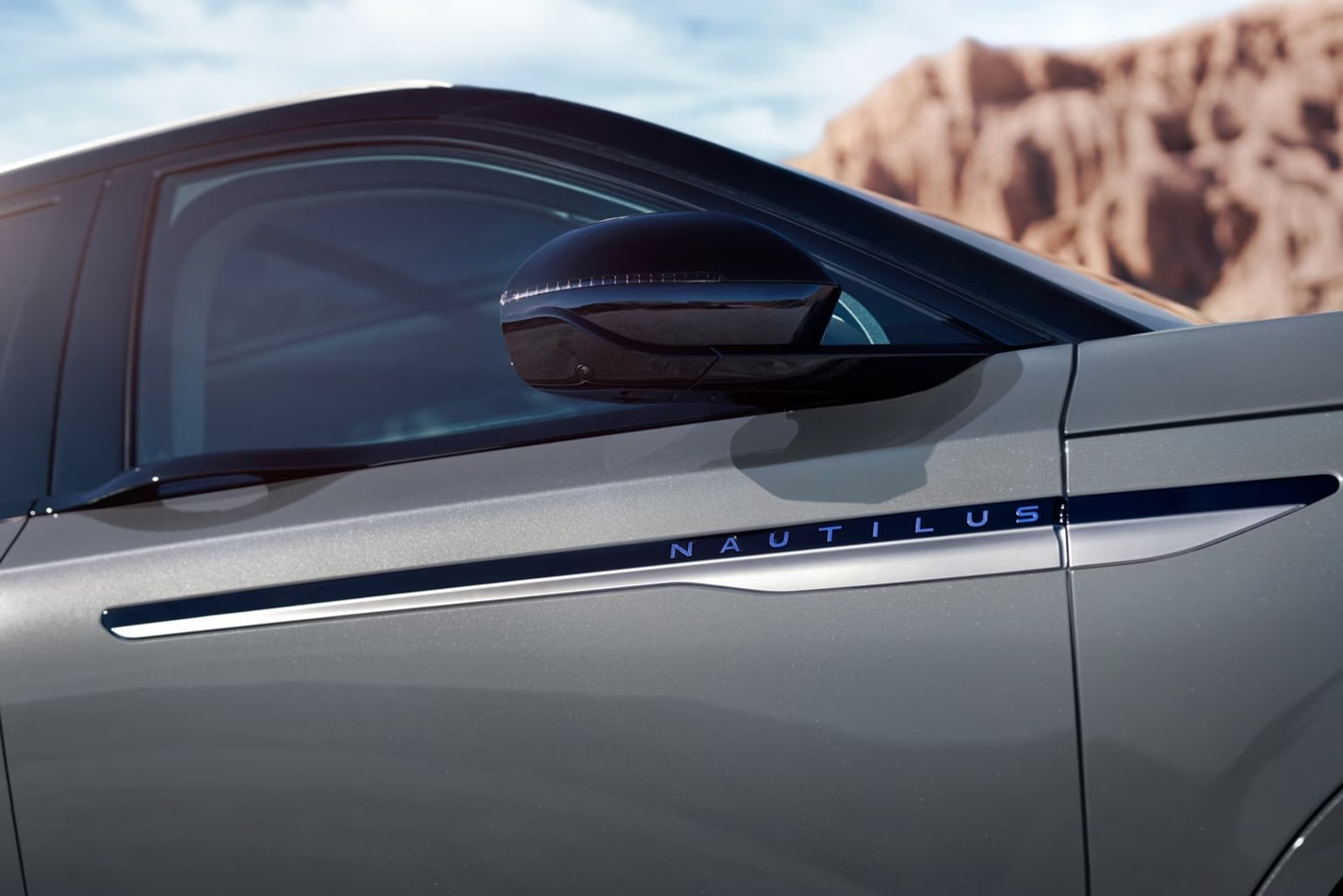 The image shows a close-up view of the side of a silver vehicle. In the background, there appears to be a rocky, mountainous landscape.
