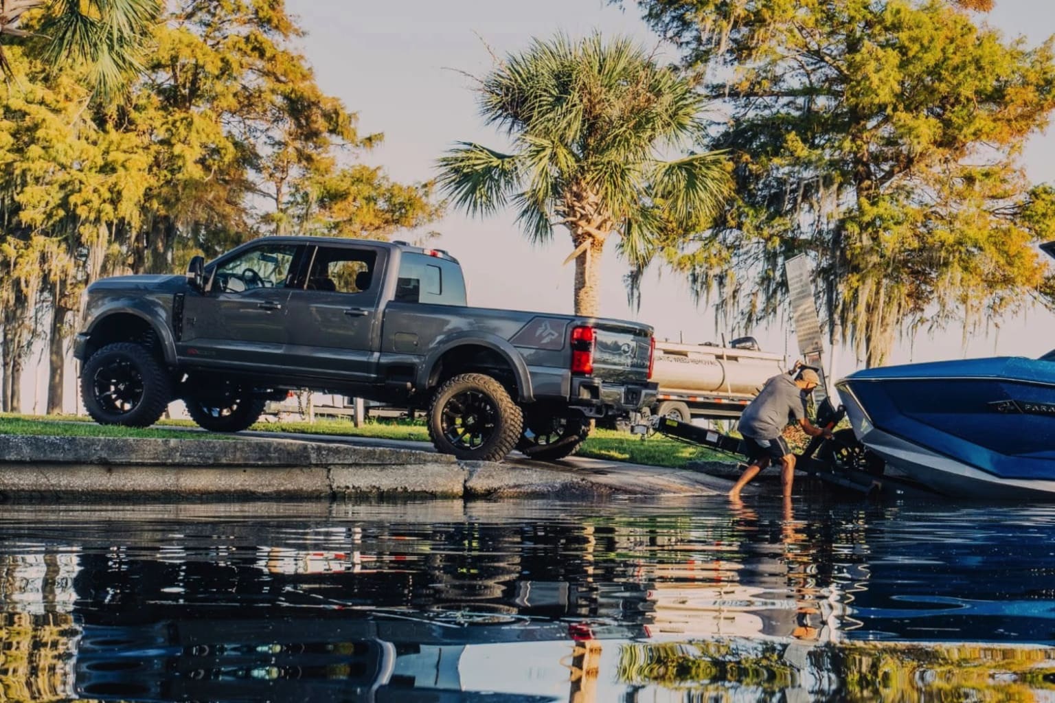 A large, lifted pickup truck parked on the edge of a body of water, with palm trees and lush vegetation in the background.