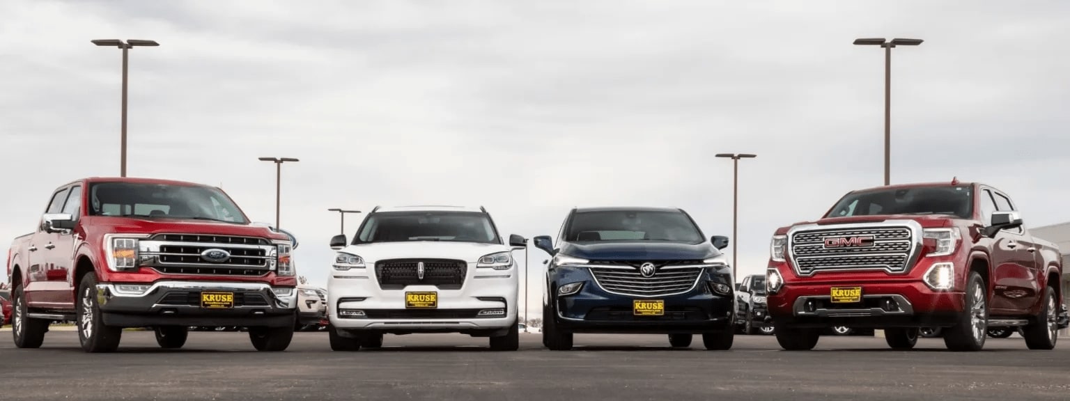 A row of large pickup trucks in various colors parked in an open area with tall lighting fixtures in the background.