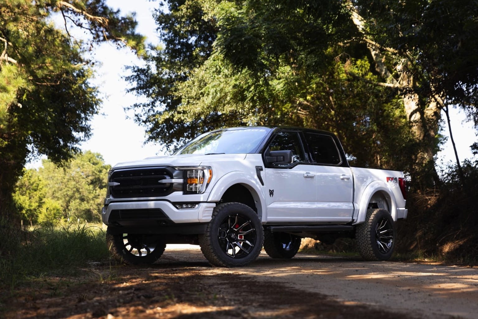 A white pickup truck with large off-road tires and a lifted suspension stands on a dirt path surrounded by lush, green foliage and trees.