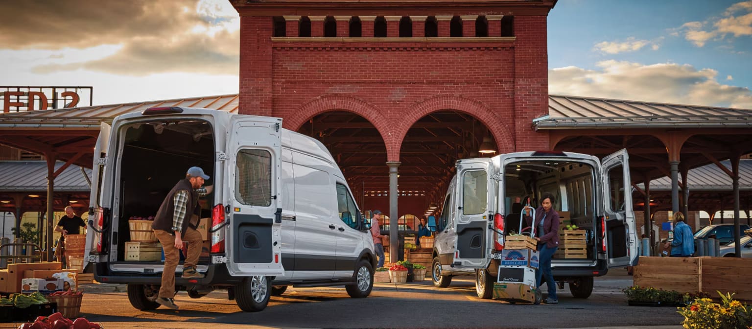 Two white vans are parked in front of a red brick building with arched openings, as people load and unload crates of produce.