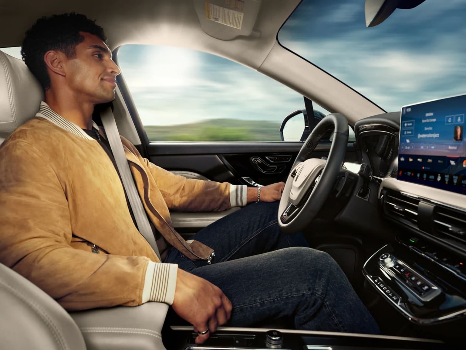 A man in a yellow jacket sits in the driver's seat of a car, with a scenic countryside landscape visible through the windshield in the background.