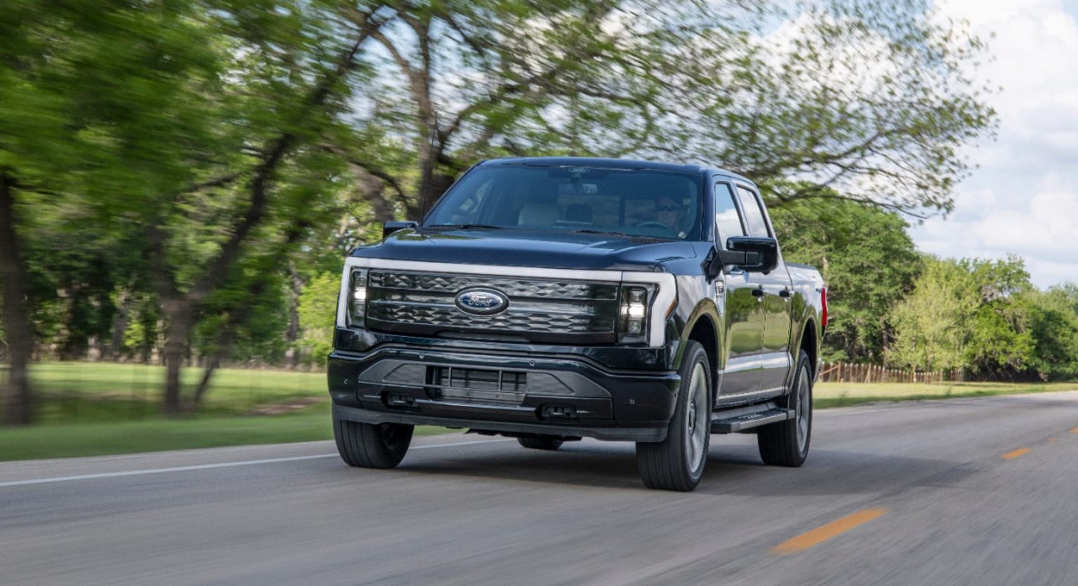 a black pickup truck driving down a road next to a lush green field with trees on both sides of the road