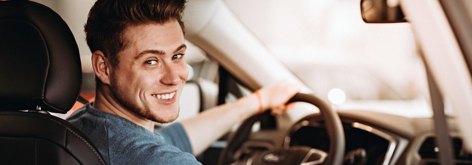 A smiling young man sitting in the driver's seat of a car, showing the interior of the vehicle in the background.