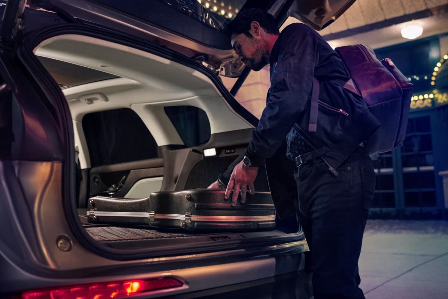 A person in a dark suit is standing in the open trunk of a vehicle, surrounded by a dimly lit indoor setting.
