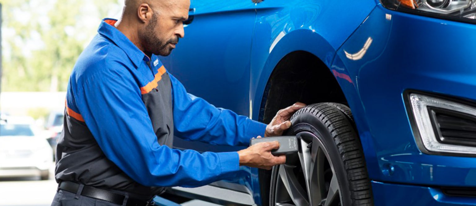 An automotive technician in a blue uniform is performing maintenance on the tire of a blue vehicle.